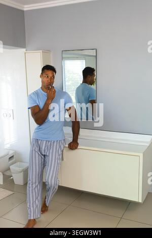 Middle-aged man standing and brushing teeth with toothbrush in bathroom, leaning on floating vanity Stock Photo