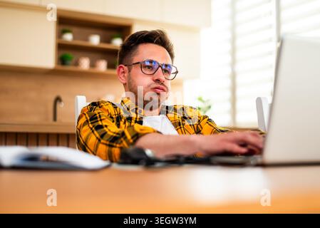 A freelancer studies something on his laptop, surrounded by a notebook and desk items in a modern home workspace. He is exhausted and worried. Stock Photo