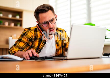 Young man is working from his home. He is using laptop and talking on smartphone. He appears worried. Stock Photo