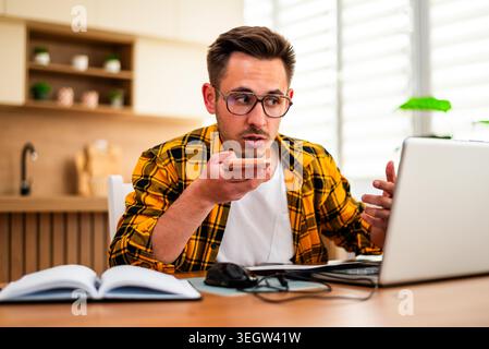 Pensive young man is working from his home. He is using laptop and talking on smartphone. Stock Photo