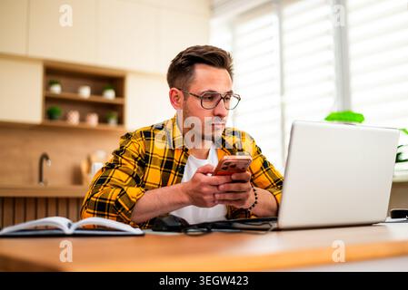 Freelance remote worker is working on his laptop from his home. He is typing on smartphone. Stock Photo