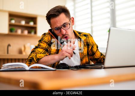 Young man is working from his home. He is using laptop and talking on smartphone. He appears angry and frustrated. Stock Photo
