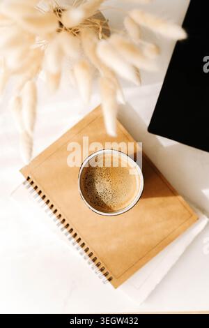 Minimal desk flat lay with coffee cup, notebook and laptop corner on a white surface, soft shadows and clean workspace aesthetic. Top view Stock Photo