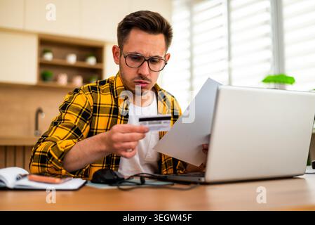 Worried young man holds a credit card while working remotely on his laptop, appearing worried and stressed. Stock Photo