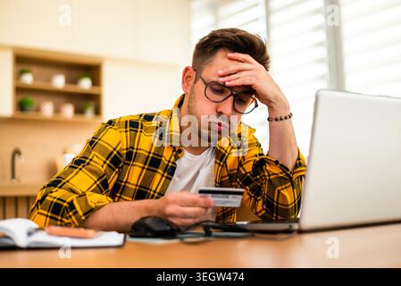 Anxious young man holds a credit card while shopping online on his laptop, appearing worried and stressed. Stock Photo