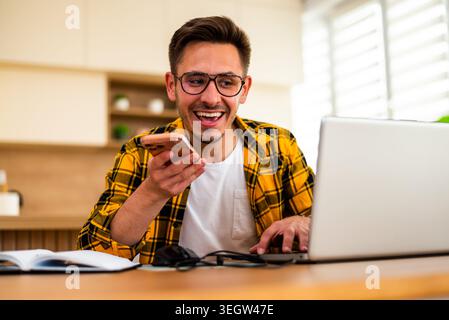 Happy young man is working from his home. He is using laptop and talking on smartphone. Stock Photo