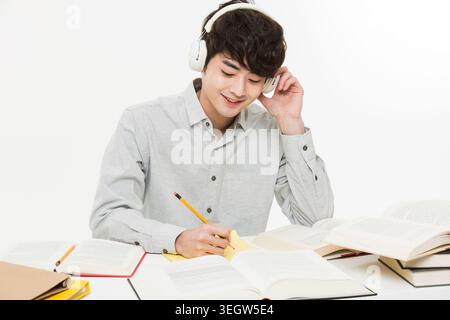A young Asian student sits at a desk, studying with a book and pencil, wearing headphones and a calm expression. Stock Photo