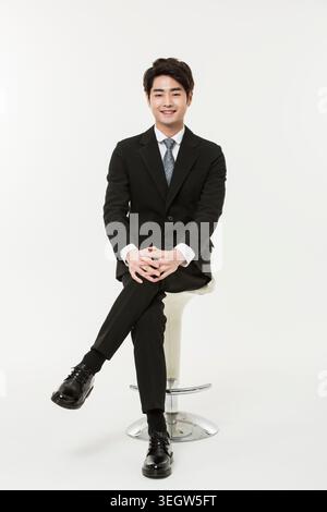 A young Asian businessman sits confidently on a stool, smiling calmly in a modern office setting. Stock Photo