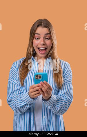Astonished young woman with european appearance holds smartphone and looks at camera in disbelief, showing dramatic surprise against a bright beige backdrop. Stock Photo
