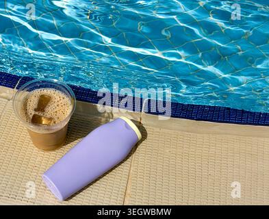 Iced coffee in a clear cup with ice cubes beside a purple water bottle on a poolside deck next to a shimmering blue swimming pool Stock Photo