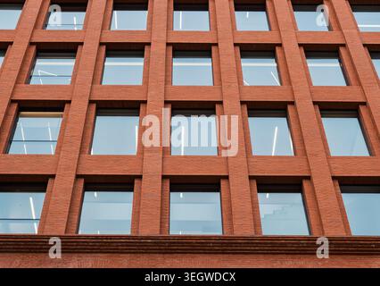 Red brick building facade showcasing a modern design with numerous square glass windows reflecting the urban daytime sky Stock Photo