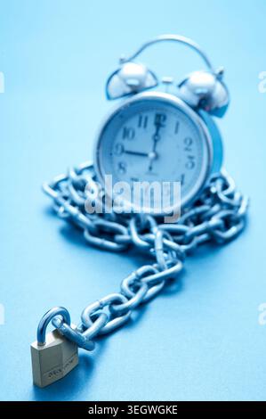 A silver alarm clock is chained to a lock on a blue background. Stock Photo