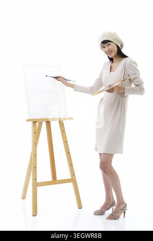 A young woman in a white dress and beret stands in front of an easel, holding a paintbrush and palette, ready to create a new painting. Stock Photo