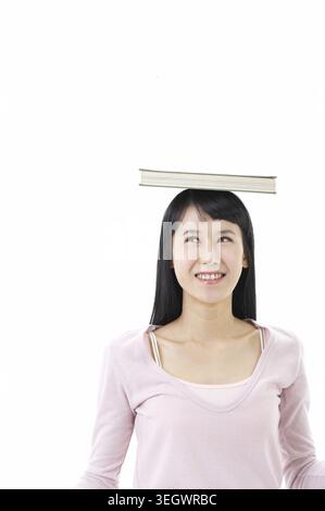 A young woman with long black hair and a pink top is balancing a book on her head. Stock Photo