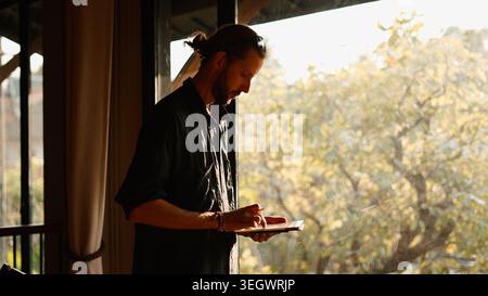 Creative man drawing on tablet near window, reflecting and working from home Stock Photo