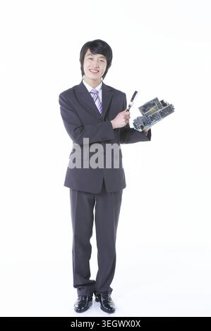 A young Asian professional in a suit holds a computer motherboard and a screwdriver, standing in a clean and modern office. Stock Photo