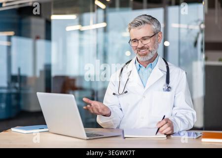 Smiling mature doctor wearing a white coat and stethoscope conducting a telemedicine session, holding a pen and taking notes at a modern office desk u Stock Photo