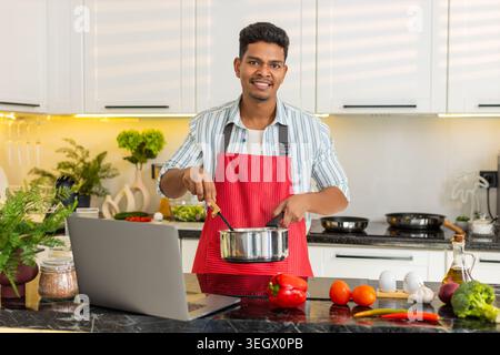 Indian man cooking healthy soup while smiling at laptop in home kitchen for weight loss meal prep. Arabian guy following online recipe for clean eating lunch with focus joy comfort during work break. Stock Photo