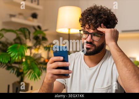 Arabian man at home checks smartphone gets bad news game loss lottery frowns shakes head no sighs. Egyptian guy on sofa reads message about result looks upset frustrated sad reacts to negative alert. Stock Photo