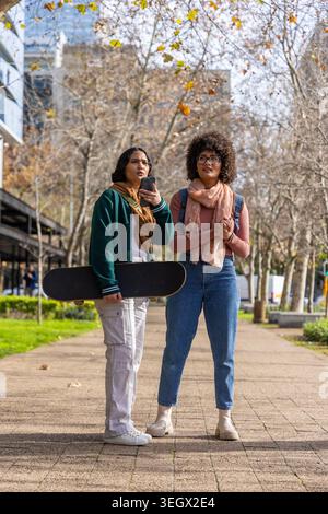 Diverse female friends standing on paved city walkway holding skateboard smartphone wearing scarves Stock Photo