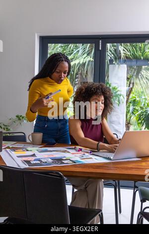 Two African American women working in studio wearing burgundy and mustard with laptop and sketches Stock Photo