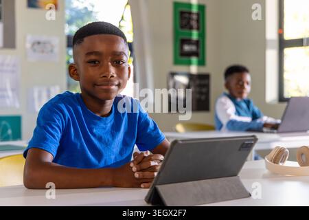 African American preteen boys sitting at white desks in classroom, using tablet and laptop Stock Photo