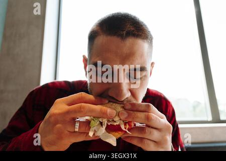 Young man eating a fresh burger near window light in modern cafe interior. Concept of fast food lifestyle comfort hunger lunch and casual urban dining Stock Photo
