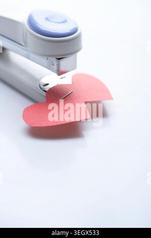 A red paper heart is being cut out of a sheet of paper by a hole punch with a metal heart-shaped punch. Stock Photo