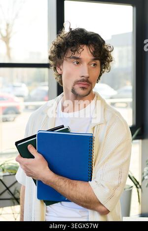 A charming young man stands in an office, holding colorful notebooks and looking thoughtful. Stock Photo