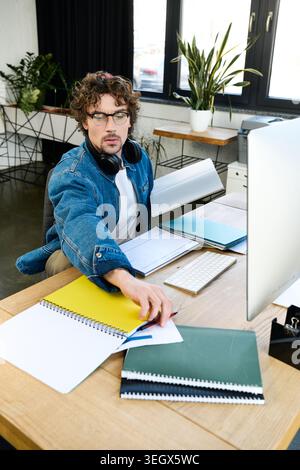 A stylish young man, wearing a denim jacket and headphones, focuses on his work in a bright office. Stock Photo