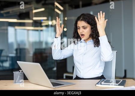 Frustrated businesswoman reacting with hands raised and mouth open, looking at a laptop screen in a modern office, expressing shock and stress from unexpected failure or technical issues Stock Photo