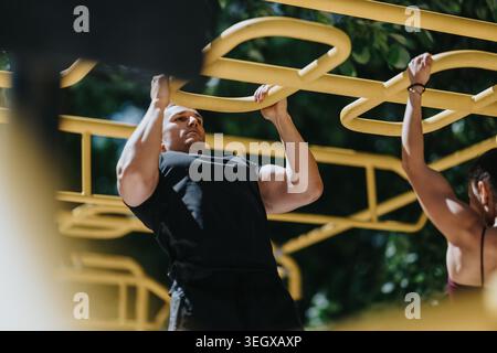 Male fitness athlete doing pull-ups on yellow outdoor gym bars with partner training Stock Photo