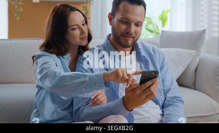 Caucasian happy smiling couple using mobile phone together looking in smartphone hugging at home carefree family man with woman pointing at screen Stock Photo