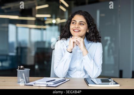 Young indian business woman sitting at her office desk, looking away and smiling, imagining future goals, career achievements, and a positive outlook for growth and progress Stock Photo