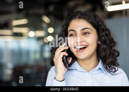 Young indian businesswoman in a light blue shirt smiling brightly while having an engaging conversation on her mobile phone in a modern office environment. Representing communication and connectivity Stock Photo
