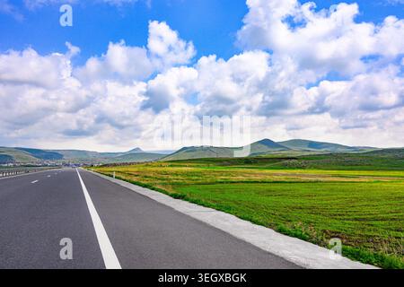 Long straight road stretches through green field under cloudy blue sky Stock Photo