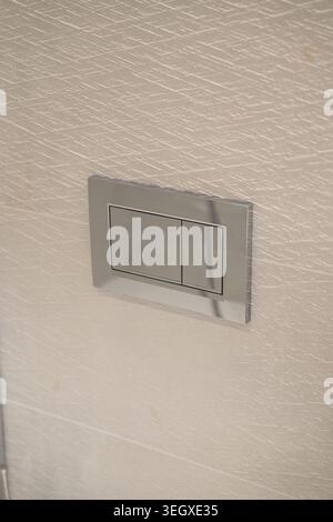 Chrome dual flush plate button installed on a textured, light-colored bathroom wall, offering water conservation and a sleek, contemporary design for Stock Photo
