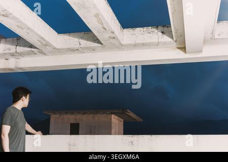 Young man standing on rooftop overlooking modern concrete structure against dramatic twilight sky Stock Photo