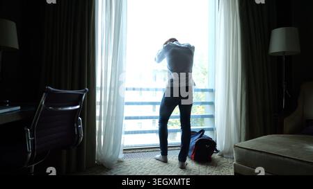 Tired young businessman in shirt standing near large window in modern hotel room or apartment interior after long trip. Exhausted manager resting Stock Photo