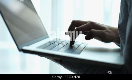 Male hand of young businessman typing text on laptop keyboard near window in modern office. Professional manager working on computer with natural Stock Photo