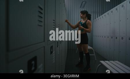 Focused Woman Prepares For Training in Locker Room, Taking Wireless Earbuds From Charging Case, Turning on Music, and Heading Workout. Fitness Lifestyle, Motivation, Technology, and Active Routine. Stock Photo