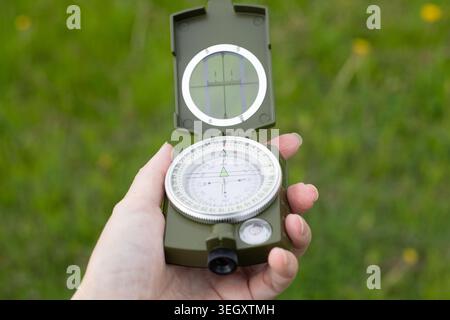 Hand holding open military magnetic compass outdoors on green grass background. Cartography gear used for hiking adventures, geography education or ta Stock Photo