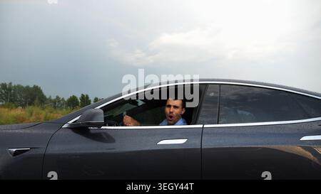 Young businessman driving on electric car in circular path on asphalt road after rain. Happy manager showing grimaces and fooling around while riding Stock Photo