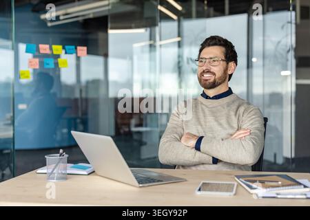 Confident businessman in casual attire smiling with arms crossed, looking away while seated at a modern glass office desk with laptop and documents, thoughtful and optimistic Stock Photo