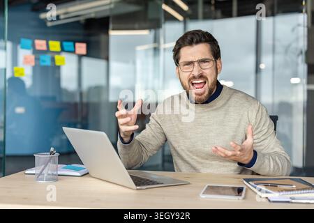 Stressed young businessman shouting and gesturing in frustration while sitting at his office desk with a laptop, reflecting work related problems and burnout Stock Photo