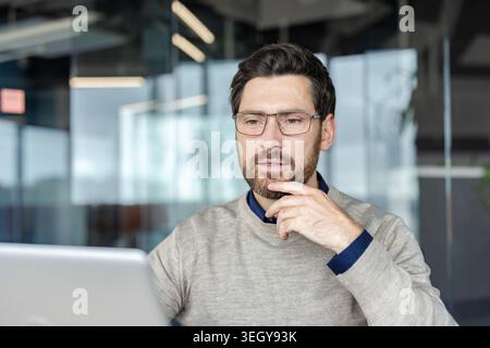 Young businessman wearing glasses and a sweater concentrating on his work at a modern office, thoughtfully touching chin while looking at a laptop screen Stock Photo