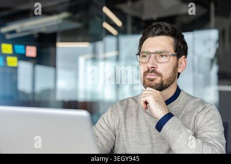 Businessman wearing glasses and a casual sweater concentrating, thinking, and planning intently while working on a laptop in a modern office environment Stock Photo