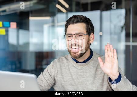 Young bearded professional man in an office wearing glasses actively participating in a video conference call, smiling and waving hand to greet the other participants Stock Photo