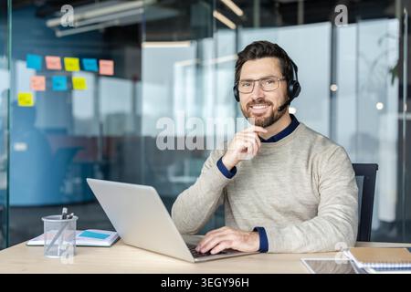 Businessman wearing a headset and spectacles is typing on a laptop, providing customer support or online consultation, while smiling and looking at the camera in a contemporary office environment Stock Photo