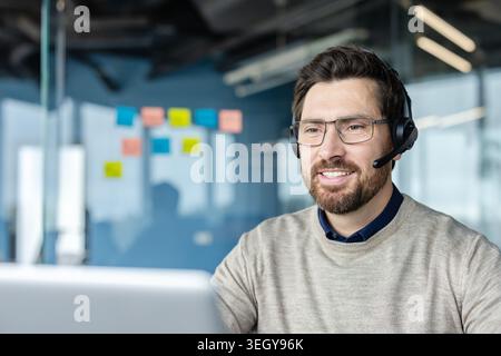 Smiling bearded man wearing eyeglasses and a headset, looking at a laptop while working as a customer support representative in a modern office environment Stock Photo